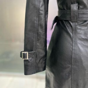 May include: A close-up of a black leather trench coat. The coat has a belt at the waist and a buckle detail on the sleeve. The leather appears smooth with a slight sheen. The coat is a classic style.