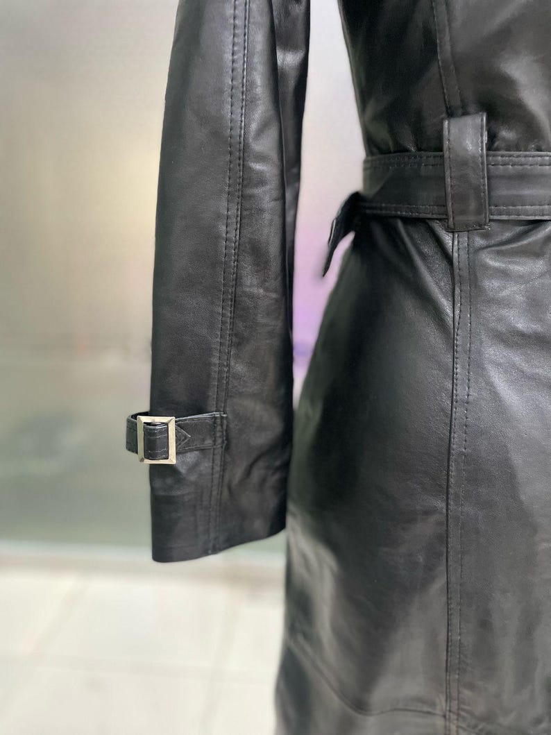 May include: A close-up of a black leather trench coat. The coat has a belt at the waist and a buckle detail on the sleeve. The leather appears smooth with a slight sheen. The coat is a classic style.
