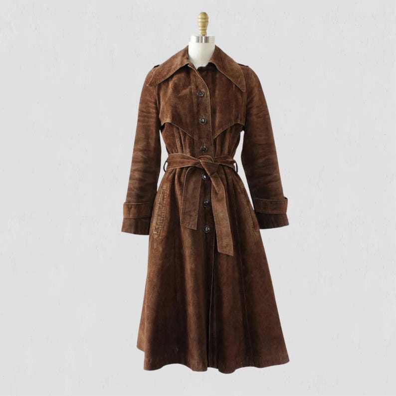 May include: A brown suede trench coat with a belted waist and button-up front. The coat has a classic collar, long sleeves, and a knee-length hem. The coat is displayed on a mannequin against a plain background.