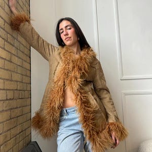 May include: A tan suede coat with brown faux fur trim on the collar and cuffs. The coat is open, revealing a glimpse of the wearer's midriff. The model is wearing light blue jeans.