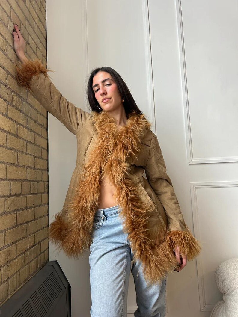 May include: A tan suede coat with brown faux fur trim on the collar and cuffs. The coat is open, revealing a glimpse of the wearer's midriff. The model is wearing light blue jeans.