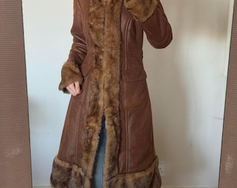 May include: A brown faux leather coat with faux fur trim. The coat features a collared neckline, long sleeves, and pockets. The faux fur trim is on the collar, cuffs, and down the front. The coat is knee-length.