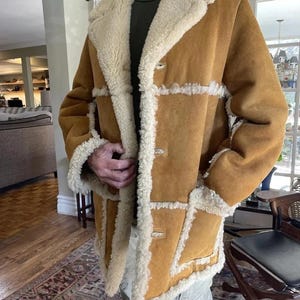 May include: A tan suede coat with cream-colored shearling lining and trim. The coat features a button-up front, large lapels, and patch detailing. The coat is knee-length and has a classic, warm aesthetic.