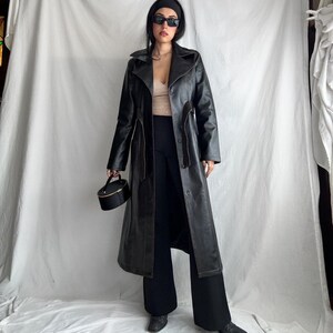 May include: A long black leather trench coat with a belt and a button closure. The coat has a collar and long sleeves. The model is wearing black pants and sunglasses.