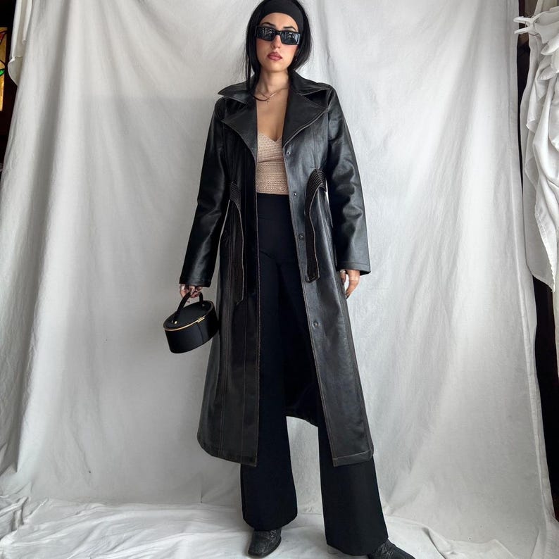 May include: A long black leather trench coat with a belt and a button closure. The coat has a collar and long sleeves. The model is wearing black pants and sunglasses.