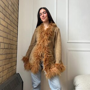 May include: A tan suede coat with brown faux fur trim around the collar, cuffs, and front. The coat has a button closure and a classic silhouette. The model is wearing light blue jeans. The coat is a vintage style.