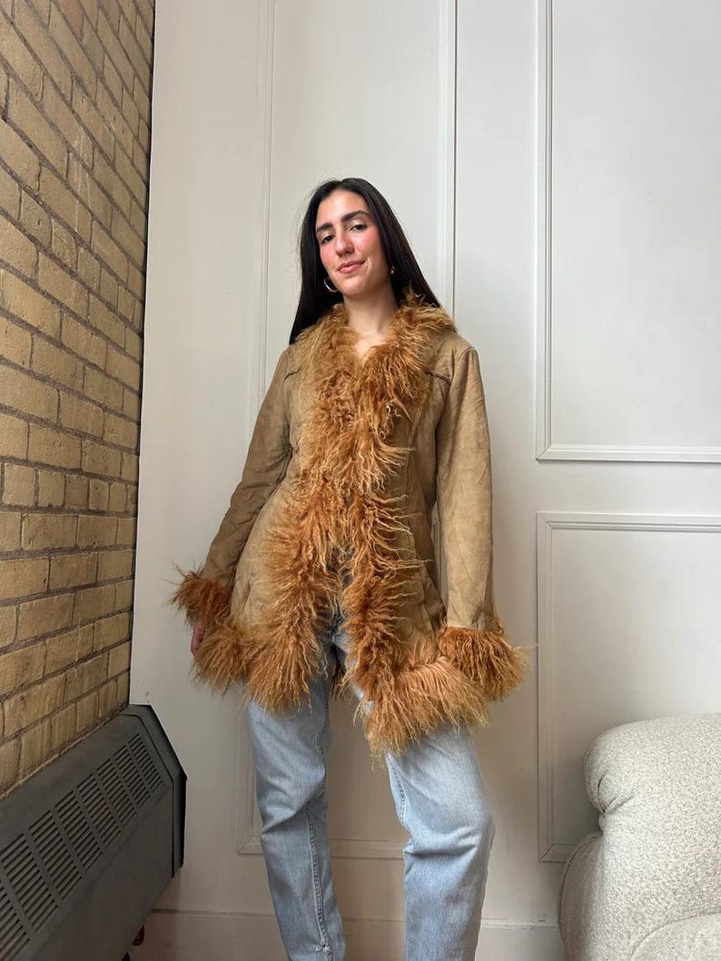 May include: A tan suede coat with brown faux fur trim around the collar, cuffs, and front. The coat has a button closure and a classic silhouette. The model is wearing light blue jeans. The coat is a vintage style.