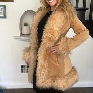 May include: A tan suede coat with a faux fur trim around the collar, cuffs, and hem. The coat has a classic design with a tailored fit and a warm, inviting aesthetic. The faux fur is a light brown color.