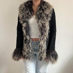 May include: A black jacket featuring a faux fur collar and cuffs. The collar and cuffs are a mix of grey, brown, and cream-coloured faux fur. The jacket has long sleeves and a suede-like texture.