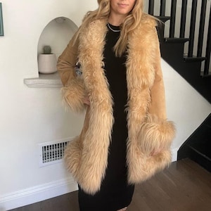 May include: A tan faux fur coat with a long, shaggy collar and cuffs. The coat is a light brown color and has a soft, suede-like texture. The coat is worn over a black dress.