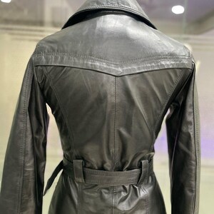 May include: A black leather trench coat with a belted waist and a notched collar. The coat has long sleeves and a classic design. The back of the coat is visible, showing the tailored fit and stitching details. The coat is hanging on a wooden hanger.