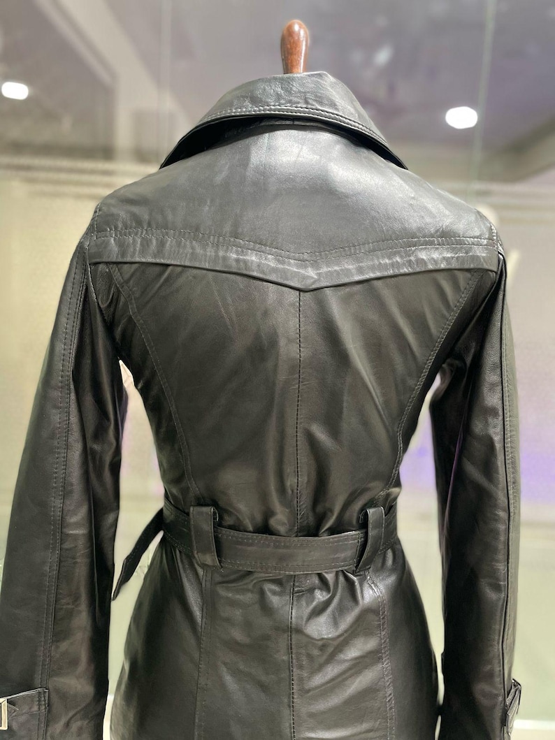 May include: A black leather trench coat with a belted waist and a notched collar. The coat has long sleeves and a classic design. The back of the coat is visible, showing the tailored fit and stitching details. The coat is hanging on a wooden hanger.