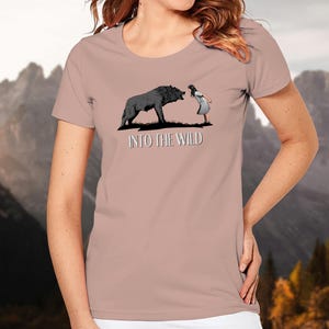 Può includere: Una maglietta rosa chiaro con un'illustrazione in bianco e nero di un lupo e una ragazza. Il testo "Into the Wild" è stampato sotto l'illustrazione.
