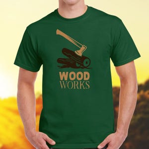Könnte beinhalten: Ein grünes T-Shirt mit einem braunen Axt- und Holzscheit-Motiv und dem Text "WOOD WORKS" auf der Vorderseite.