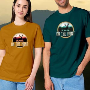 May include: Mustard yellow and teal green t-shirts with a circular graphic featuring a deer silhouette, trees, and the text "ON THE HUNT". The shirts are short-sleeved and worn by two people, with a mountain backdrop.