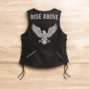 Può includere: Gilet in pelle nera con un'aquila ricamata bianca e la scritta "RISE ABOVE". Il gilet ha lacci laterali e la scritta "ZAYN INDUSTRIES". Il gilet è esposto su una superficie di legno.