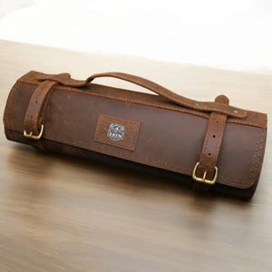 Peut inclure: Un rouleau à couteaux en cuir marron avec une sangle et une fermeture à boucle. Le rouleau porte un logo "ZAYN".
