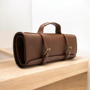 以下が含まれることがあります： ハンドルと2つのバックルが付いた茶色の革製のロールアップケース。ケースは閉じており、木製の表面の上に置かれています。