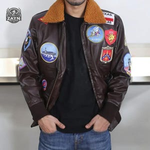 May include: A brown leather bomber jacket with a faux fur collar and multiple patches. The patches feature various designs, including airplanes, ships, and military insignia. The jacket has a zipper closure and two pockets.