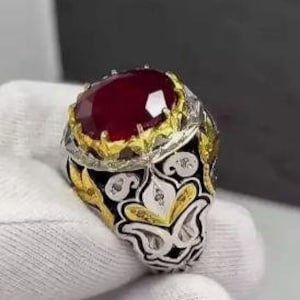 May include: A silver ring with a large, oval-cut, deep red gemstone. The setting features gold accents and intricate filigree detailing. Small, clear stones are set around the band.