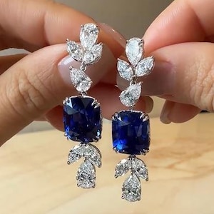 May include: A pair of drop earrings featuring a large, cushion-cut deep blue gemstone at the center. Each earring is accented with multiple pear-shaped and marquise-cut clear stones arranged in a leaf-like pattern above and below the blue center.