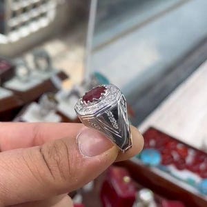 May include: A silver ring featuring a large, oval, red gemstone, encircled by smaller, clear stones. The ring's band has an intricate, geometric pattern. The ring is held in a person's hand.