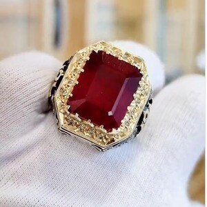 Op de afbeelding: Een goudkleurige ring met een grote, rechthoekige, dieprode edelsteen. De edelsteen is gezet in een gedetailleerde, sierlijke bezel. De ring heeft een vintage of antieke stijl, met ingewikkelde details rond de band.