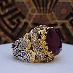 Puede incluir: Un anillo con una gran piedra preciosa de color rojo oscuro y corte ovalado, engastada en un marco dorado rodeado de pequeñas piedras claras. La banda es de metal plateado con intrincados grabados y pequeñas piedras verdes incrustadas.