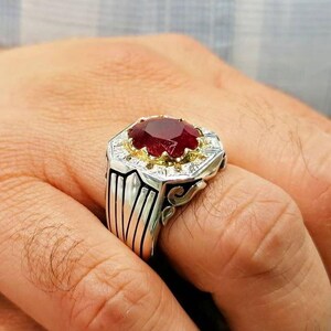 May include: A silver ring with a large, oval-cut ruby gemstone. The ruby is set in a gold-colored bezel, surrounded by smaller white gemstones. The ring has a detailed, geometric design on the band.