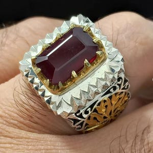 Könnte beinhalten: Ein silberner Ring mit einem großen, rechteckigen, tiefroten Edelstein. Der Edelstein ist in einer goldfarbenen Lünette gefasst. Der Ring hat ein detailliertes, strukturiertes Design mit silbernen und goldenen Akzenten.