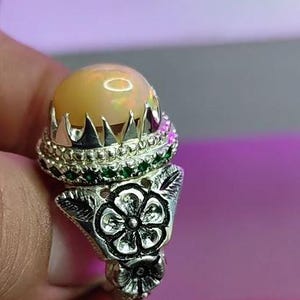 May include: A silver ring featuring a large, round, light-yellow opal gemstone. The ring has a floral design on the band and is accented with green and pink gemstones. The opal is set in a crown-like setting.