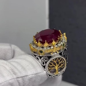 May include: An elaborate ring with a large, oval, deep red gemstone. The ring has a gold crown setting with small diamonds and green accents. The band is silver with a gold tree of life design.