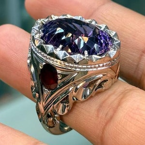 May include: A silver-toned ring featuring a large, oval-cut purple gemstone set in a pronged bezel. The band displays intricate metalwork with a smaller, oval-cut dark red gemstone embedded on the side, surrounded by decorative scroll-like details.