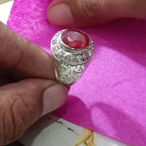 Puede incluir: Un anillo de plata con una gran piedra preciosa de color rojo intenso y corte ovalado en el centro, rodeada por un halo de pequeñas piedras claras de corte redondo. La banda de plata presenta intrincados grabados florales en los laterales.