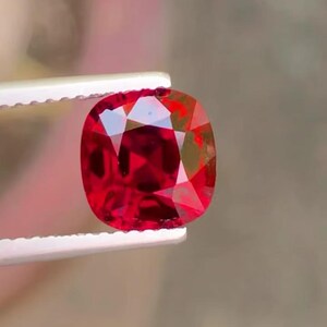 Pode incluir: Uma pedra preciosa vermelha facetada, com corte almofada, é segurada por pinças brancas. A gema tem um alto grau de clareza e brilho, refletindo a luz. A pedra preciosa é de uma cor vermelha profunda e saturada, com inclusões visíveis.