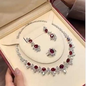 May include: A jewelry set featuring a necklace, earrings, and a ring, presented in a red box. The set includes a silver-toned metal with red gemstones and clear crystals. The necklace and earrings have a floral design.