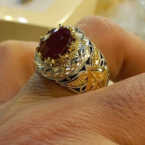 Puede incluir: Un anillo dorado con una gran piedra preciosa roja oscura de talla ovalada. El anillo presenta intrincados diseños de filigrana y estrellas en la banda y alrededor del engaste de la piedra preciosa. El anillo se lleva en un dedo.