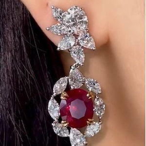 May include: A drop earring featuring a large, round, faceted deep red gemstone at the center, surrounded by a halo of marquise and pear-shaped clear white stones. A cluster of similar clear white stones forms the top section of the earring.