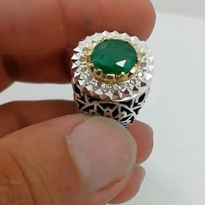 May include: A silver ring with a large, oval-cut emerald gemstone at the center. The emerald is surrounded by a halo of small, clear gemstones and a gold setting. The ring has an intricate, openwork design on the band.