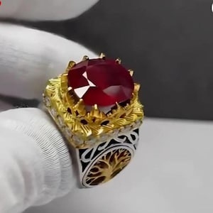 Puede incluir: Un anillo ornamentado con una gran gema facetada de color rojo intenso. El anillo tiene un engaste dorado con detalles intrincados y una banda plateada con un diseño de árbol de la vida. La gema es el punto focal.