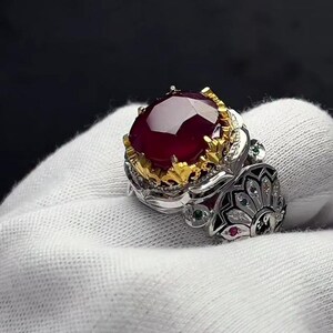 Puede incluir: Un anillo ornamentado con una gran gema de color rojo oscuro, probablemente un rubí, engastada en una banda de oro y plata. El anillo presenta detalles intrincados, incluyendo piedras de acento más pequeñas y un diseño decorativo en el lateral.