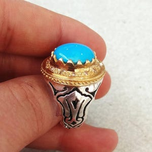 May include: A silver ring with a large, oval, bright blue gemstone. The ring has a gold-colored bezel around the stone and a detailed silver band with an ornate design. The ring is a statement piece.