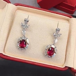 May include: A pair of elegant drop earrings displayed in a red jewelry box. Each earring features a large, oval, red gemstone surrounded by smaller, clear stones. The earrings have a delicate, dangling design with additional clear stones.