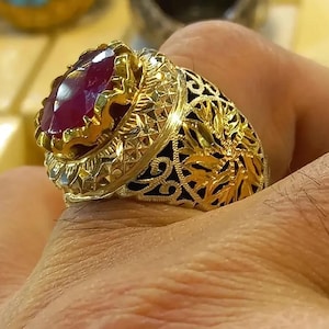 May include: A gold-colored ring featuring an oval, deep red gemstone. The ring has intricate filigree and floral patterns, with a dark background enhancing the gold's shine. The gemstone is set in a detailed bezel.