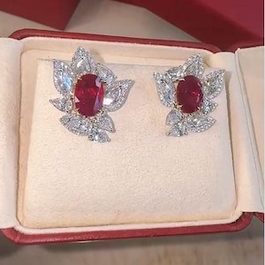 May include: A pair of stud earrings featuring a central oval-cut deep red ruby surrounded by a cluster of marquise and round-cut clear white stones arranged in a floral-like pattern, set in a metallic frame and presented in a red jewelry box.