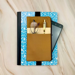 May include: A composition notebook with a blue and white splatter design, a brown pocket, and a black elastic band. The pocket holds eyeglasses, a gold pen, and a tablet.