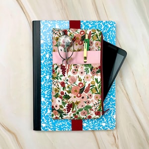 May include: A floral fabric notebook cover with a pink pocket, holding eyeglasses and a pen, rests on a composition notebook. A tablet is tucked inside the cover. The cover features a red elastic band and a floral pattern with pink, red, and green flowers.