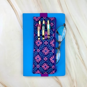 May include: A blue notebook with a floral pen case and eyeglasses. The pen case is navy blue with a pink and blue daisy pattern. Three gold pens are in the pen case. The eyeglasses have a silver frame.