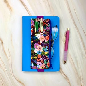 May include: A blue notebook with a floral patterned pencil pouch attached to the front. The pouch has a zipper closure and is holding three pens and a pair of glasses. A pink pen is lying on the table next to the notebook.