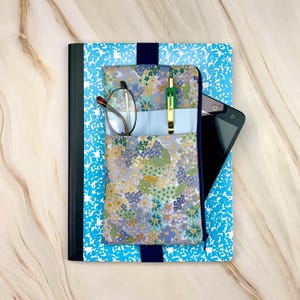 May include: A blue and white speckled composition notebook with a dark blue elastic band holding a floral fabric pouch. The pouch contains a pair of metal-rimmed glasses and a green pen, with a black tablet partially tucked behind it.